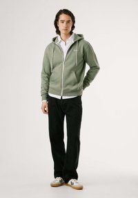 Sudadera verde con cremallera y capucha ajustable, con bolsillos frontales y puños acanalados, combinada con una camisa blanca de cuello y pantalones negros.