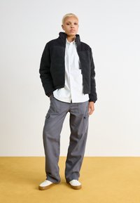 Veste en polaire noire avec un col en velours côtelé, chemise rayée blanche, pantalon cargo gris et baskets blanches avec semelles marron. Design simple et texturé.