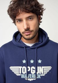 Felpa blu navy con una grafica frontale che presenta "TOP GUN MAVERICK", stelle e dettagli a forma di ali; tessuto testurizzato e cappuccio con cordino.