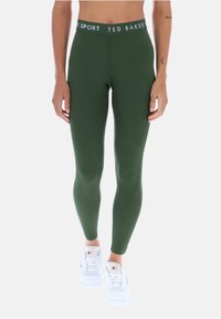 Zelené legíny vyrobené z hladkého a elastického materiálu, s bielym elastickým pásom s logom. Skombinované s bielymi teniskami.