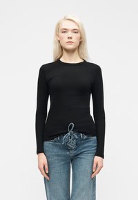 Γυναίκα με μακριά ξανθά μαλλιά, που φοράει εφαρμοστή μαύρη ριγέ μπλούζα με μακριά μανίκια και ψηλόμεσα μπλε τζιν με λεπτομέρεια κορδονιών μπροστά.