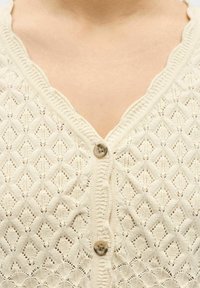 Cardigan en maille crème avec motif en losange et deux boutons marron fermés sur la poitrine du torse supérieur d'une personne.