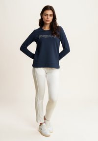 Sudadera de manga larga de color navy con el texto brillante "FREDDY", combinada con pantalones blancos ajustados y zapatillas blancas. Tejido suave y corte relajado.