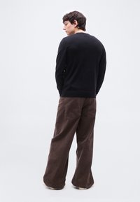 Homme portant une chemise noire à manches longues et un pantalon large marron, debout, légèrement tourné vers la gauche sur un fond blanc uni.