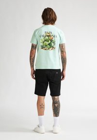 T-shirt vert menthe clair avec un graphisme de cactus et le texte "BAJA CALIFORNIA MV" au dos, porté avec un short noir et des chaussettes blanches.