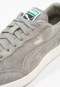 Baskets Puma Liga en daim gris avec lacets gris, semelle blanche et logo Puma vert et noir sur la languette.