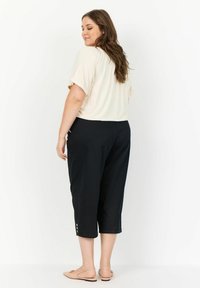 Ljusbeige kortärmad topp kombinerad med svarta capribyxor med knappdetaljer vid nederkanten. Modellen står med ryggen mot kameran.