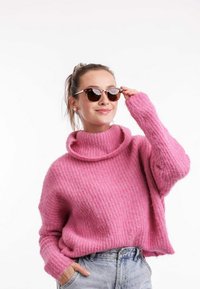 Růžový pletený svetr s vysokým límcem a žebrovanou strukturou, spárovaný s kulatými slunečními brýlemi a světle modrými džínovými šortkami.