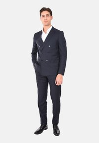 Completo blu navy a doppio petto con bottoni argentati, fitting su misura, abbinato a una camicia bianca e scarpe nere lucide, in piedi su uno sfondo neutro.