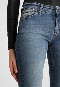 Osoba ubrana w jasnoniebieskie, wyblakłe dżinsy i czarny, dopasowany top z długim rękawem, ręka spoczywająca przy boku.