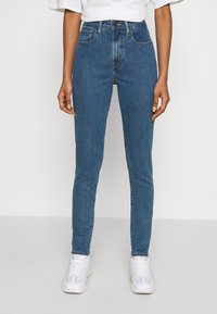 Vaqueros de tiro alto en azul con un corte slim, que cuentan con cinco bolsillos y un desvanecimiento sutil. Combinados con zapatillas blancas y una camiseta blanca holgada.