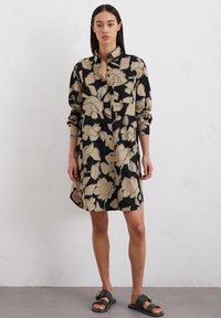 Vestito camicia nero con motivo floreale beige, con bottoni sul davanti e maniche lunghe. Indossato con sandali neri con una fascia larga e fibbia dorata.