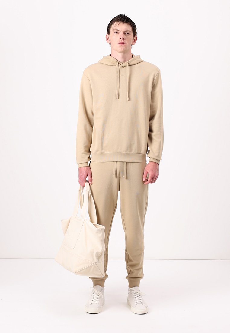 Sweat-shirt beige et joggeurs assortis en tissu doux, avec une capuche à cordon, une coupe décontractée et des poches latérales. Portant un sac fourre-tout beige clair.