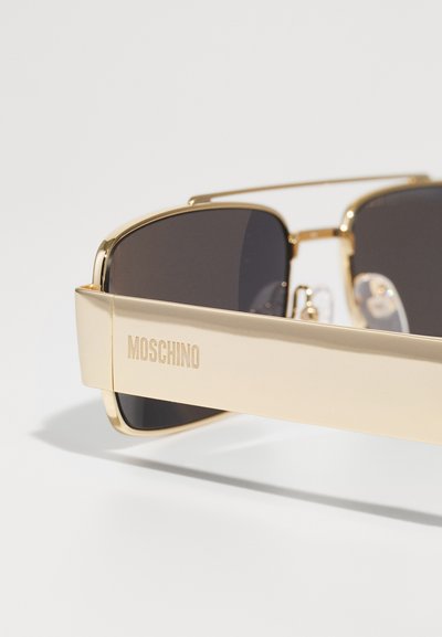Sluneční brýle ve zlatém tónu s tmavými zrcadlovými skly. Mají silný rám a nožičky s vyrytým názvem značky "MOSCHINO" na boku.