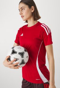 Camiseta atlética roja con acentos blancos, textura de tela, mangas cortas y logo. Sosteniendo un balón de fútbol blanco y negro con detalles estampados.