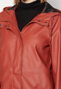 Rode waterdichte jas met een matte afwerking, voorzien van een ritssluiting aan de voorkant, een capuchon en drukknopen. De kraag heeft een contrasterende zwarte voering.