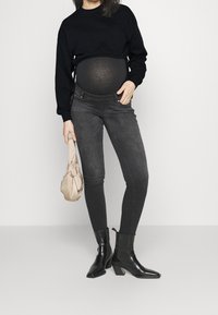 Maglietta nera a maniche lunghe con vita arricciata sopra un pancione visibile, abbinata a jeans skinny scuri e stivaletti neri lucidi.