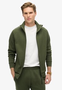 Olivengrøn zip-up jakke med høj krave, ribbede ærmer og sidelommer, parret med en hvid t-shirt. Glat, blød stoftekstur.