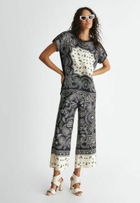Top a righe nere e crema con pantaloni a gamba larga caratterizzati da motivi paisley. L'outfit include sandali con tacco spesso e occhiali da sole bianchi oversize.