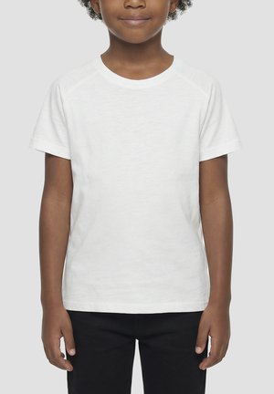 Enfant aux cheveux bouclés portant un t-shirt blanc uni à manches courtes et un pantalon noir, debout devant un fond gris clair.