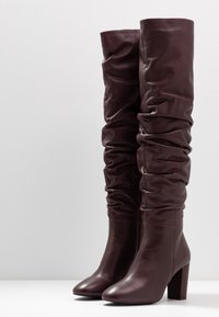 Botas de cuero burdeos hasta la rodilla con un diseño holgado, tacones apilados y textura suave; costuras notables a lo largo de las uniones.