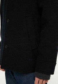 Mörk marinblå jacka i ullblandning med två svarta knappar, sidoficka och texturerade ärmar. Funktioner inkluderar en skördkant och figursydd design.