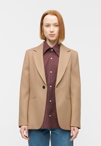 Μπεζ σακάκι με διαμορφωμένους ώμους, ένα μαύρο κουμπί, συνδυασμένο με πουκάμισο με μπορντό ρίγες και μπλε τζιν. Λεία υφή υφάσματος.