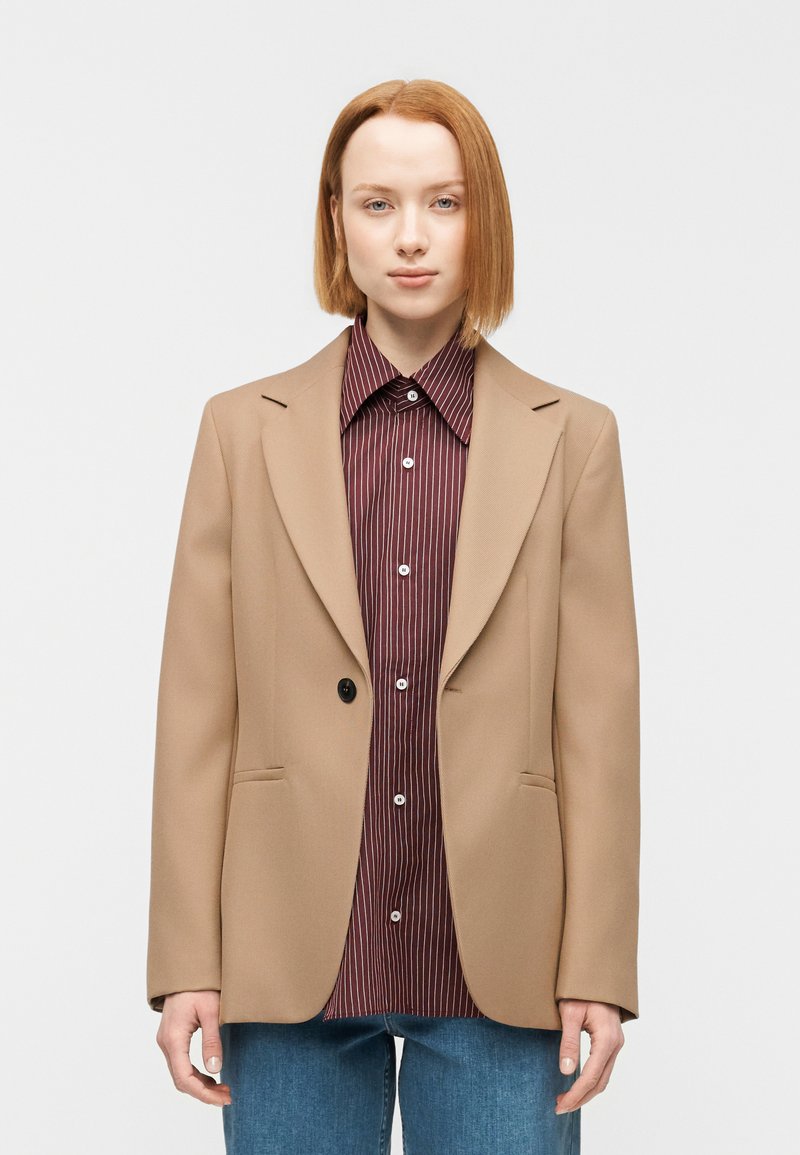 Μπεζ σακάκι με διαμορφωμένους ώμους, ένα μαύρο κουμπί, συνδυασμένο με πουκάμισο με μπορντό ρίγες και μπλε τζιν. Λεία υφή υφάσματος.