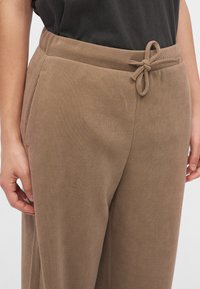 Gros plan d’une personne portant un pantalon côtelé beige avec une taille à cordon noué et un haut noir, montrant une coupe décontractée et une poche latérale.