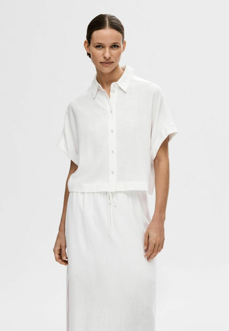 Chemise blanche courte à boutons et à manches courtes, en tissu léger et texturé. Associée à une jupe longue à cordon de serrage assortie.
