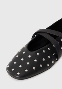 Scarpa piatta nera con borchie rotonde argentate e cinturini incrociati sulla parte superiore, esposta su uno sfondo bianco.
