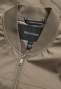 Jachetă bomber de culoare maro deschis, cu guler ribbed, fermoar metalic și etichetă interioară cu sigla "Matinique" și dimensiunea "L". Textură fină.