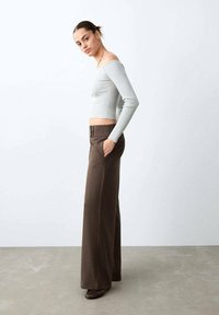 Žena stojící s rukama v kapse hnědých širokých kalhot, oblečená v světle šedém žebrovaném topu s dlouhým rukávem spadajícím z ramen a hnědých botách.