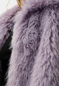 Lilla kunstpels med en lang, shaggy tekstur. Har et sort underlag og subtile knapdetaljer på siden. Blødt og fluffy udseende.