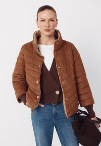 Brun quiltet jakke med høj krave, tanforing, trykknapper og ribstrikkede manchetter, stylet med en mørkebrun cardigan og blå jeans.