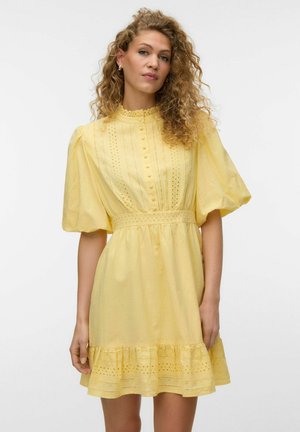 Femme aux cheveux bouclés portant une robe jaune à œillets avec des manches bouffantes, un devant boutonné et un ourlet à volants, debout devant un fond uni.