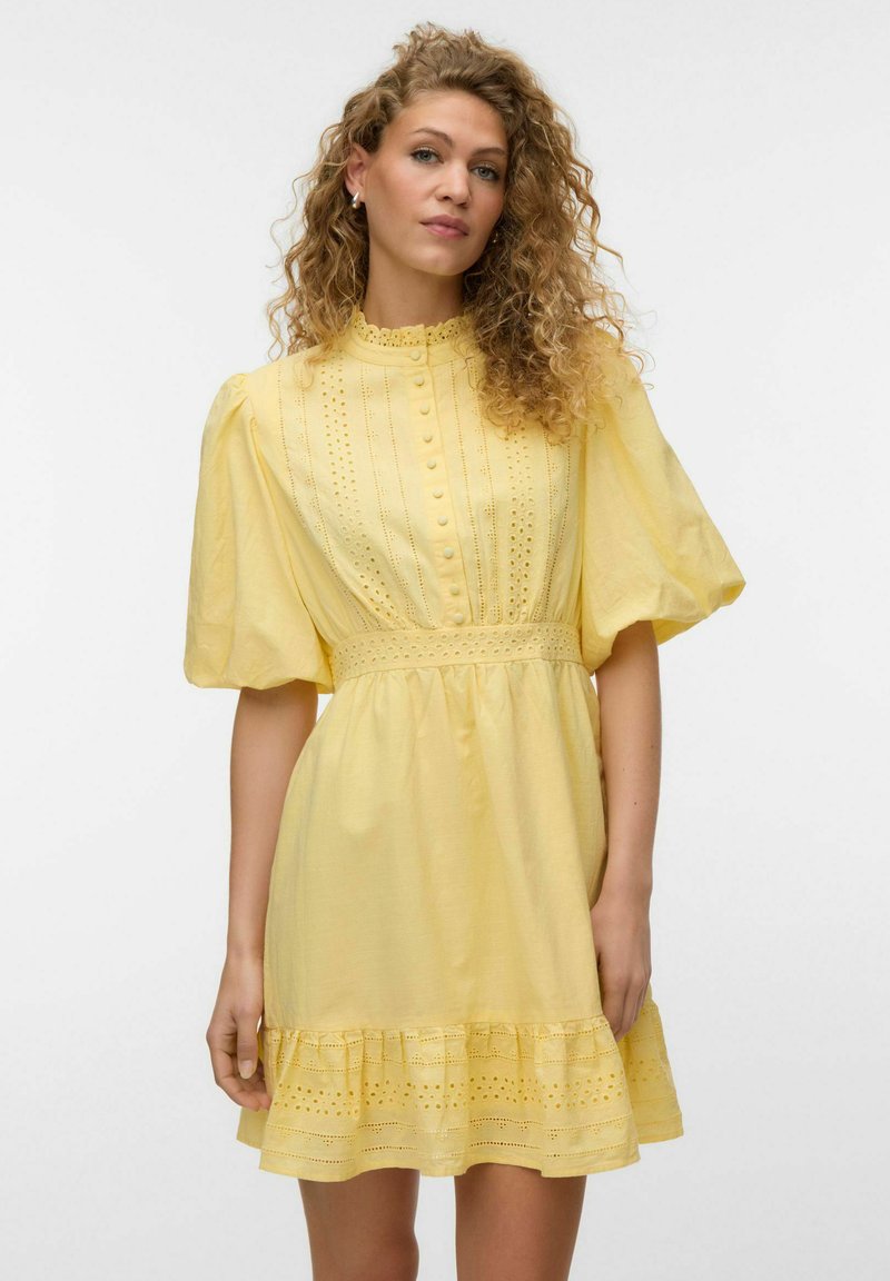 Femme aux cheveux bouclés portant une robe jaune à œillets avec des manches bouffantes, un devant boutonné et un ourlet à volants, debout devant un fond uni.