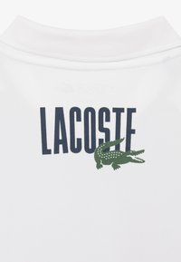 Polo bianco con logo "LACOSTE" blu navy e emblema di coccodrillo verde sul retro, realizzato in tessuto traspirante.