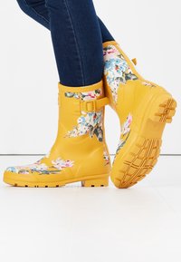 Botas de lluvia de goma amarillas con patrones florales en rosa, azul y verde. Presentan un detalle de hebilla en la parte superior y una suela texturizada para mayor agarre.