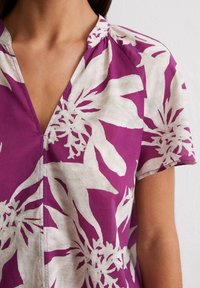 Kurzärmlige Bluse mit V-Ausschnitt in Dunkelpink mit einem weißen Blumenmuster. Hergestellt aus leichtem Stoff mit einer glatten Textur und dezentem Glanz.