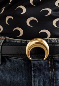 Cinto de pele sintética preta com uma fivela em forma de lua crescente dourada, usado por cima de jeans de ganga com uma blusa preta com padrões de luas crescentes.