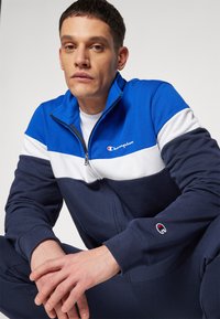 Modra in temno modra zip-up jakna z belimi vodoravnimi črtami, iz mešanice bombaža, vezeno logo Champion na prsih in rokavu, teksturirani manšeti.