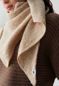 Beige gebreide sjaal met een zachte textuur en driehoekige vorm, om de nek gedrapeerd, met een klein logo-label aan de zijkant.
