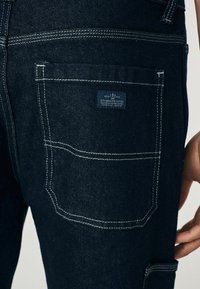 Donkerdenim jeans met witte stiksels, voorzien van een logo patch op de achterzak en een gestructureerd ontwerp met meerdere zakken.