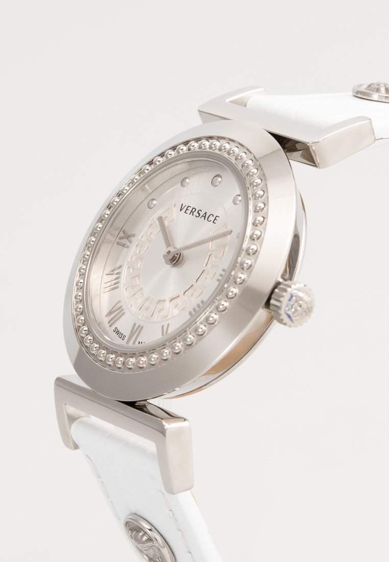 Montre ronde en ton argent avec un bracelet en cuir blanc. Présente un cadran argenté texturé, des détails en perles autour de la lunette et des chiffres romains comme indicateurs.