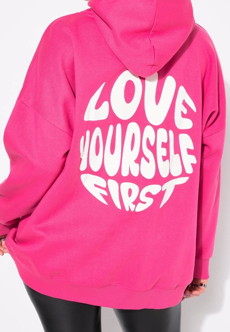 Personne portant un sweat à capuche rose vif avec un texte blanc en gras "LOVE YOURSELF FIRST" dans le dos, associé à un pantalon noir.