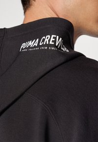 Sudadera negra con un acabado texturizado. Presenta un logo blanco de "PUMA CREW" en la parte superior de la espalda. El diseño incluye una capucha y costuras cosidas.