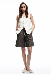 Weißes, ärmelloses Vest mit drei Knöpfen, kombiniert mit braunen, knielangen Shorts mit elastischem Bund und Seitentaschen.