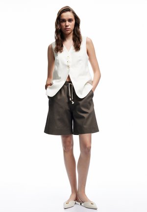 Gilet blanc sans manches avec trois boutons, associé à un short marron à longueur genou avec une taille élastique et des poches latérales.