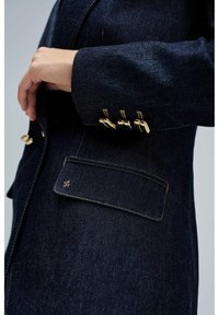 Chaqueta de mezclilla oscura con un corte ajustado. Presenta detalles de botones dorados en los puños, costuras sutiles y un bolsillo lateral con un detalle de logo.